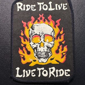 Könnte beinhalten: Schwarzer rechteckiger Aufnäher mit einem weißen Totenkopf, der von orangefarbenen und gelben Flammen umgeben ist. Der Text "RIDE TO LIVE" steht über dem Totenkopf und "LIVE TO RIDE" darunter. Der Aufnäher hat einen dunkelblauen Rand.