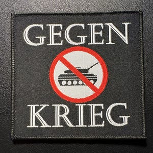 Könnte beinhalten: Ein schwarzer quadratischer Aufnäher mit weiß gesticktem Text „GEGEN KRIEG“. Ein roter Kreis mit einem Strich durch eine Panzergrafik befindet sich in der Mitte. Der Aufnäher ist wahrscheinlich für Kleidung oder Accessoires.