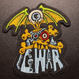 K&ouml;nnte beinhalten: Gestickter Aufn&auml;her mit einem Skelett-Design, einem Auge, Fl&uuml;geln und dem Wort "GWAR" in Blau. Der Aufn&auml;her hat gelbe, gr&uuml;ne, rote und wei&szlig;e Akzente auf schwarzem Hintergrund. Wahrscheinlich ein Band-Aufn&auml;her oder ein Merchandise-Artikel.