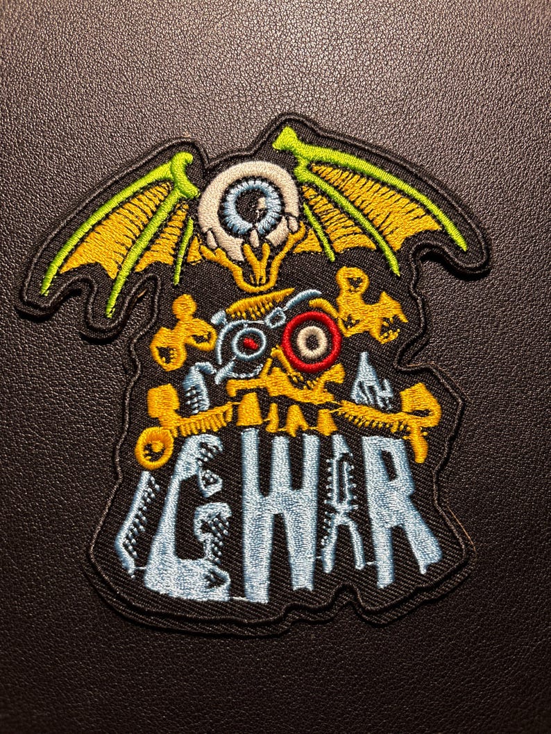K&ouml;nnte beinhalten: Gestickter Aufn&auml;her mit einem Skelett-Design, einem Auge, Fl&uuml;geln und dem Wort "GWAR" in Blau. Der Aufn&auml;her hat gelbe, gr&uuml;ne, rote und wei&szlig;e Akzente auf schwarzem Hintergrund. Wahrscheinlich ein Band-Aufn&auml;her oder ein Merchandise-Artikel.