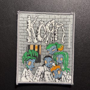 May include: Embroidered patch featuring the band name "KoRn" in white, stylized lettering. The patch depicts a group of cartoon figures with blue faces and green or orange hair, set against a gray brick wall background.