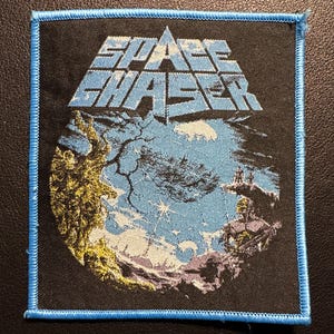 Könnte beinhalten: Quadratischer schwarzer Aufnäher mit blauem Rand. Der Aufnäher zeigt den Text "SPACE CHASER" in einer stilisierten Schriftart. Er zeigt eine Himmelszene mit einem Planeten, Sternen und Figuren in Blau-, Gelb- und Lilatönen.