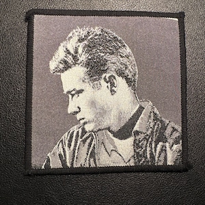 Könnte beinhalten: Quadratischer Aufnäher mit einem Schwarz-Weiß-Porträt eines Mannes im Profil. Das Bild hat eine gewebte Textur und ist von einem schwarzen Rand umgeben. Der Mann trägt ein Hemd mit Kragen und eine Jacke.