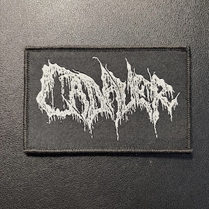 以下が含まれることがあります： 黒い長方形の布製ワッペン。白の刺繍で「CADAVER」という文字が、ギザギザとした滴るようなスタイルで描かれています。ワッペンにはステッチ入りの縁取りがあり、衣類やアクセサリーに取り付けて装飾として使用するように設計されています。