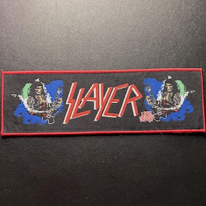Könnte beinhalten: Schwarzer rechteckiger Aufnäher mit dem Wort "SLAYER" in roten und weißen Blockbuchstaben. Der Aufnäher hat einen roten Rand und zeigt zwei Illustrationen von Figuren mit Gitarren auf beiden Seiten des Wortes.
