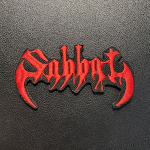 Pode incluir: Um emblema de tecido com a palavra Sabbat bordada numa fonte vermelha estilizada e irregular. As letras têm uma estética afiada e escorrida, com extremidades alongadas e curvas na primeira e na última letra, criando um design marcante.