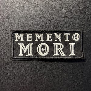 Op de afbeelding: Een rechthoekige zwarte geborduurde patch met de witte tekst MEMENTO MORI in een schreeflettertype. De patch heeft een zwarte gestikte rand en is ontworpen voor bevestiging op stoffen oppervlakken, met duidelijke witte letters op een donkere achtergrond.