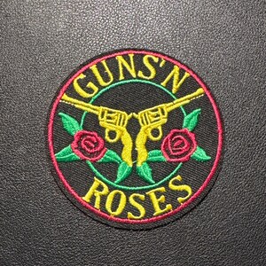 Könnte beinhalten: Runder Aufnäher mit dem Text "GUNS'N ROSES" in Gelb, der sich oben und unten wölbt. Zwei gelbe Revolver sind in der Mitte gekreuzt, mit roten Rosen und grünen Blättern.