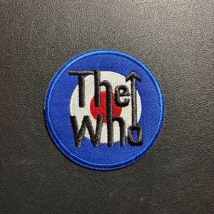 Może przedstawiać: Okrągła, haftowana naszywka z logo tarczy zespołu The Who. Projekt ma niebieską obwódkę, biało-czerwony wzór tarczy i nazwę zespołu w czarno-białych literach. Kultowy symbol.