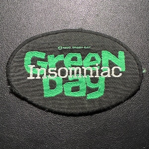 Könnte beinhalten: Ovaler Aufnäher mit den Worten "Green Day Insomniac" in grüner und weißer Schrift. Der Aufnäher hat einen schwarzen Hintergrund und einen schwarzen Rand. Der Text "©1995 Green Day" ist ebenfalls sichtbar.