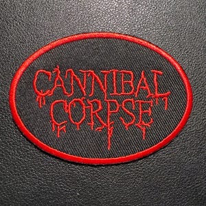 May include: Oval patch with the text "CANNIBAL CORPSE" in red, stylized lettering. The text is on a black background, framed by a red border. The patch is likely for clothing or accessories.