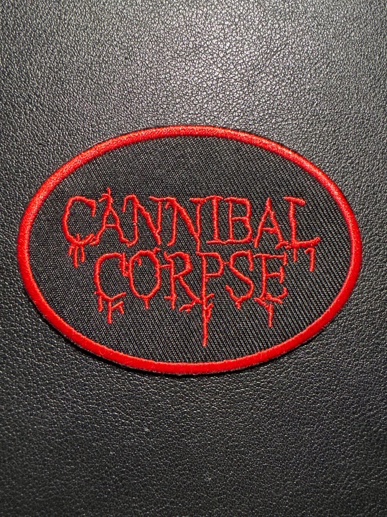 May include: Oval patch with the text "CANNIBAL CORPSE" in red, stylized lettering. The text is on a black background, framed by a red border. The patch is likely for clothing or accessories.