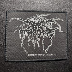 Op de afbeelding: Een zwart rechthoekig embleem met een wit, gestileerd ontwerp, mogelijk een bandlogo. De tekst "©1999 DARK THRONE U.L.T. RAZAMATAZ" is in het wit onderaan het embleem gedrukt.