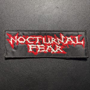 Könnte beinhalten: Ein rechteckiger, bestickter Aufnäher mit schwarzem Hintergrund. Der Text NOCTURNAL FEAR ist in Weiß mit einer gezackten, abgenutzten Schriftart gestickt. Die Buchstaben sind mit rotem Garn umrandet und akzentuiert, was einen Kontrast bildet.
