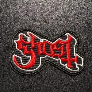Könnte beinhalten: Ein schwarzer Aufnäher mit dem Wort "Ghost" in roter gotischer Schrift, weiß umrandet. Der Aufnäher hat eine rechteckige Form mit abgerundeten Ecken und liegt auf einem strukturierten schwarzen Hintergrund.