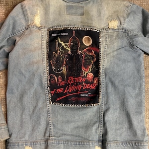 Könnte beinhalten: Hellblaue Jeansjacke mit Used-Look-Design. Auf der Rückseite befindet sich ein schwarzer Aufnäher mit einer Grafik aus dem Film "The Return of the Living Dead", umrahmt von silbernen Nieten. Auf dem Aufnäher steht "They're Back!" und "They're Hungry."