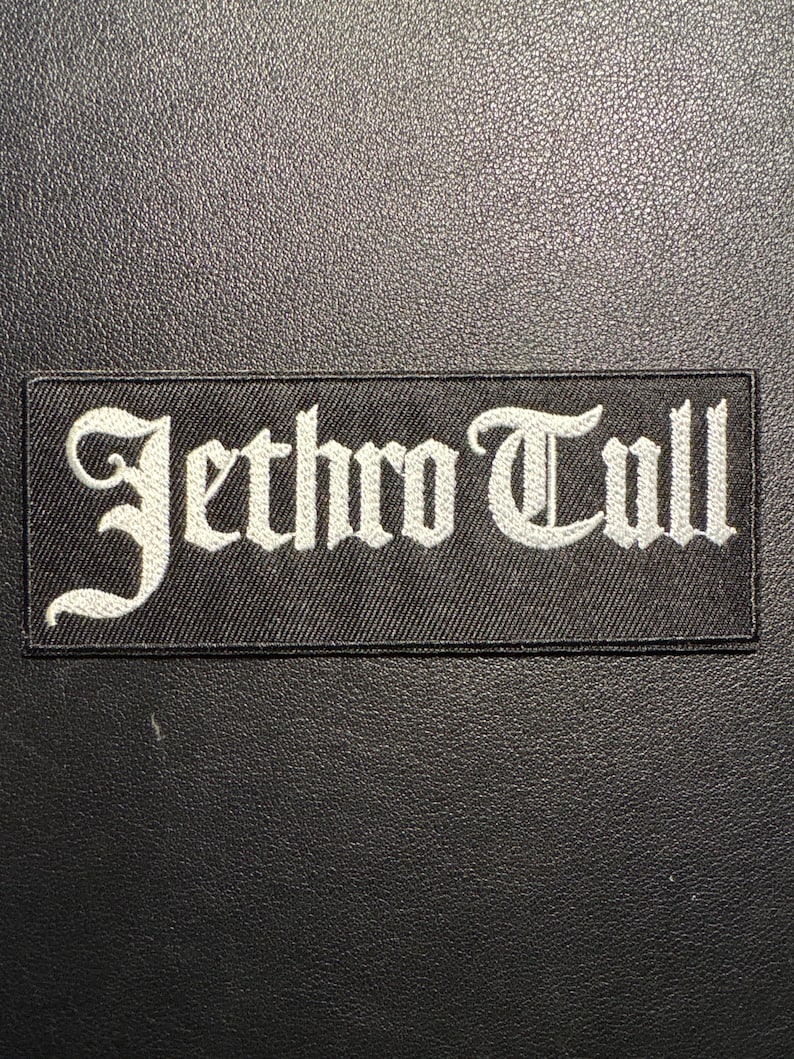 May include: A black rectangular patch featuring the embroidered name "Jethro Tull" in white, gothic font. The patch is designed for application on clothing or accessories, set against a textured black surface.