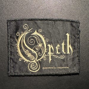 May include: Black rectangular patch with the word "Opeth" in a gothic font, and an ornate gold design. The text "©2001 OPETH U.L.T. RAZAMATAZ" is below the word Opeth. The patch is on a black textured surface.