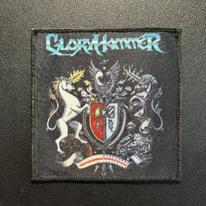 Könnte beinhalten: Ein quadratischer schwarzer Stoffaufnäher mit dem Schriftzug Gloryhammer in Blau und Weiß am oberen Rand. Das Design zeigt ein weißes Einhorn auf der linken Seite, einen zentralen Schild in Rot und Blau mit Blitzen sowie eine geflügelte Figur.