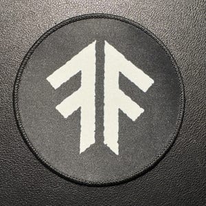 May include: A circular black patch featuring a white arrow symbol. The patch has a stitched edge and is displayed on a dark surface. The arrow design is the central focus.