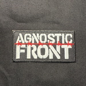 Puede incluir: Parche rectangular negro con las palabras "AGNOSTIC FRONT" en letras blancas. Diseño de alambre de púas rojo bordado encima de la palabra "FRONT". El parche está sobre un fondo de tela negra.