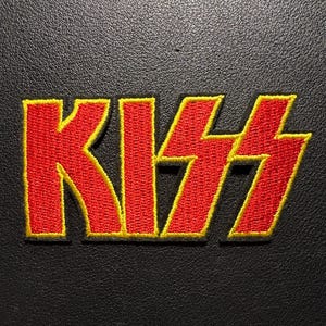 Peut inclure: Écusson brodé avec le mot "KISS" en rouge vif et un contour jaune. Le patch est posé sur un fond noir texturé, créant un contraste saisissant. Idéal pour personnaliser vêtements et accessoires.