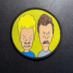 Op de afbeelding: Geborduurde ronde patch met twee stripfiguren met overdreven gezichtsuitdrukkingen. Eén figuur heeft geel haar en een blauw shirt, de ander heeft bruin haar en een grijs shirt. De patch heeft een gele achtergrond en een zwarte rand.