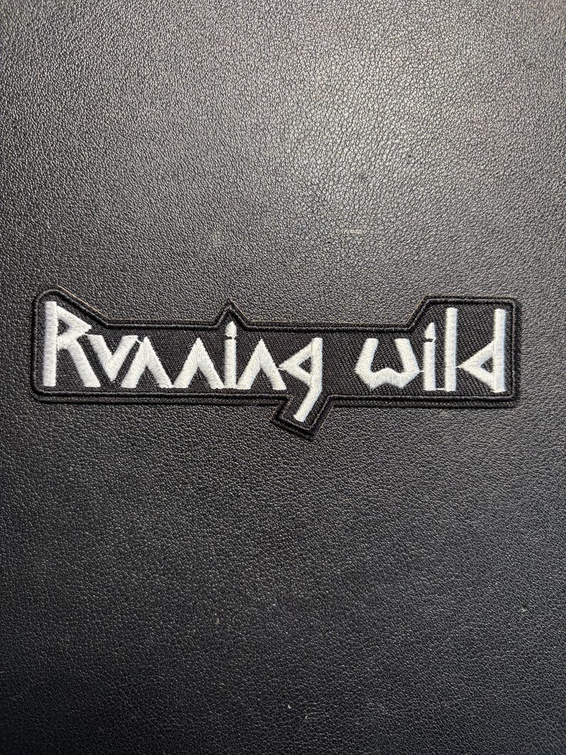 May include: Black embroidered patch featuring the words "Running Wild" in white, with a distinctive font. The patch has a black border and is set against a textured black surface.