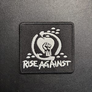 Könnte beinhalten: Schwarzer quadratischer Aufnäher mit weiß gesticktem Design und Text. Das Design zeigt eine geballte Faust in einem Kreis mit einem Apfel und einem Pfeil, darunter die Worte "RISE AGAINST".