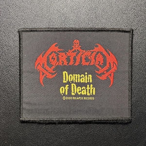 Puede incluir: Un parche rectangular de tela negra con texto bordado en rojo que dice Mortician con un pequeño icono de calavera encima. Debajo, el texto bordado en amarillo dice Domain of Death, seguido del texto 2000 Relapse Records.