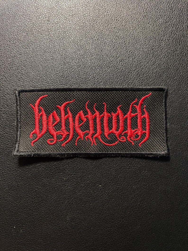 K&ouml;nnte beinhalten: Ein rechteckiger, schwarz bestickter Aufn&auml;her mit dem Wort behemoth in einer stilisierten roten Schriftart. Der Aufn&auml;her hat eine strukturierte Stoffbasis mit einem schwarzen Rand und ist zur Anbringung an Kleidung oder Textilien gedacht.