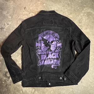 Op de afbeelding: Zwarte spijkerjas met een paarse afbeelding op de rug. De afbeelding bevat de tekst "LORD OF THIS WORLD" en "BLACK SABBATH" met een vleermuisillustratie. De jas heeft knoopsluitingen en zakken.