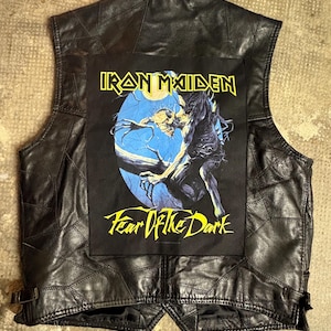 Peut inclure: Gilet en cuir noir avec un motif patchwork. Il présente un grand écusson avec le texte "IRON MAIDEN" en jaune et un graphisme d'une figure squelettique sur fond bleu. Le texte "Fear Of The Dark" est également visible.