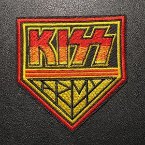 May include: Embroidered patch featuring the band name "KISS" in red and yellow block letters, with a yellow and red diamond shape below that reads "ARMY". The patch has a black border and is set against a black background.