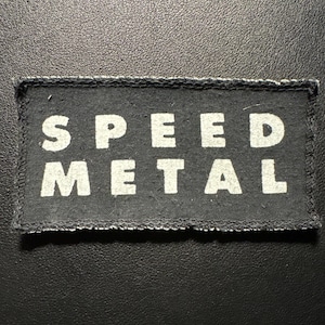 Könnte beinhalten: Schwarzer rechteckiger Aufnäher mit den Worten "SPEED METAL" in fetten weißen Großbuchstaben. Der Aufnäher hat einen genähten Rand und ist vor einem dunklen Hintergrund platziert.