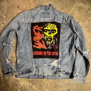 May include: Light blue denim jacket with a distressed design. The back features a black patch with the band name "Slayer" and the album title "Seasons in the Abyss" with a yellow skull graphic.