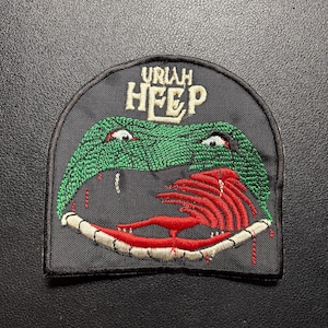 May include: A black embroidered patch featuring the text URIAH HEEP in cream letters above a green snake head with red markings, a white jaw, and a red tongue. The patch has a rounded top edge and a straight bottom edge with a black stitched border.
