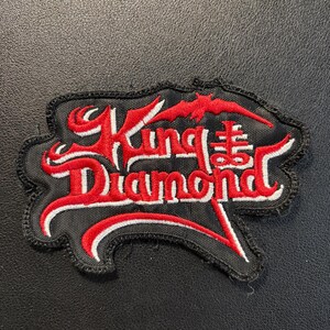 May include: A black embroidered patch featuring the text King Diamond in stylized red lettering with white outlines. Above the text is a small red bat silhouette, and to the right is a red cross symbol. The patch has a black stitched border.