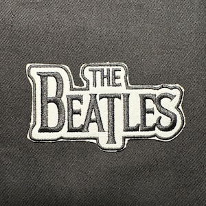 Könnte beinhalten: Gestickter Aufnäher mit dem Schriftzug "THE BEATLES" in Schwarz und Weiß. Der Aufnäher hat einen weißen Rand und ist auf einem dunkelgrauen Stoff platziert. Ideal für Jacken, Taschen oder andere Textilien.