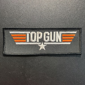 May include: A rectangular embroidered patch with a black background featuring the white text TOP GUN. The design includes horizontal orange and white stripes on both sides of the text and a white star centered below the word GUN.