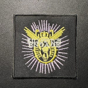 Könnte beinhalten: Ein schwarzer quadratischer Aufnäher mit einem gestickten Design. Das Design zeigt einen Totenkopf mit Krone, Flügeln und den Worten "KILLSWITCH ENGAGE" in Gelb. Der Totenkopf ist von gelben und weißen Strahlen umgeben.
