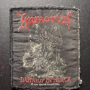 May include: Black embroidered patch featuring the word "Immortal" in red gothic lettering above a faded black and white image. The text "DAMNED IN BLACK" is below the image, also in red. The patch is square with frayed edges.
