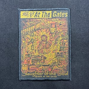 Op de afbeelding: Een rechthoekige patch met de tekst "At The Gates" bovenaan en "Slaughter of the Soul" onderaan. De patch heeft een gedetailleerde illustratie in tinten oranje, geel en bruin, met een zwarte rand. Het jaar 2019 is ook zichtbaar.