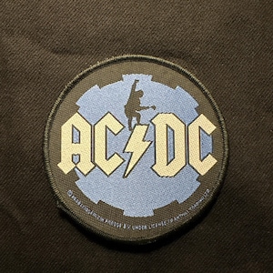 Könnte beinhalten: Runder Aufnäher mit dem AC/DC-Logo in weißen Blockbuchstaben und einem Blitz. Das Logo befindet sich auf einem hellblauen Hintergrund mit einer schwarzen Silhouette einer Figur. Der Aufnäher hat einen schwarzen Rand.