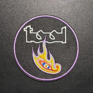Könnte beinhalten: Gestickter Aufnäher mit dem Bandnamen "TOOL" in Weiß, umrandet von einem lila Rand. Der Aufnäher zeigt ein stilisiertes Auge in einem Flammenmuster mit gelben, orangefarbenen und blauen Farben. Der Aufnäher befindet sich auf schwarzem Hintergrund.