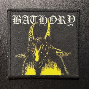 Könnte beinhalten: Ein quadratischer schwarzer Stoffaufnäher mit einer gelben Grafik einer gehörnten Ziege mit roten Augen. Das Wort BATHORY ist oben in einer weißen gotischen Schrift gedruckt. Der Aufnäher hat einen schwarzen gestickten Rand und eine Webstruktur.