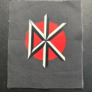 Könnte beinhalten: Ein quadratischer Stoffaufnäher in Dunkelgrau mit einem zentralen Grafikdesign. Das Motiv besteht aus einem roten Kreis, der mit einem stilisierten weiß-schwarzen geometrischen Logo überlagert ist, das an die Buchstaben DK erinnert.