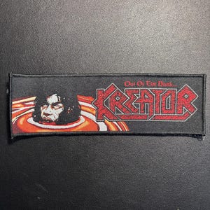 Puede incluir: Un parche rectangular negro con el nombre de la banda "KREATOR" en letras rojas. El parche presenta una ilustración de una cara en un diseño arremolinado naranja y blanco, con el texto "Out Of The Dark".