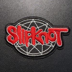 May include: Embroidered patch featuring the word "Slipknot" in red, outlined in white, on a black background. The text is in a stylized font, with a white web design behind the text. The patch has a black border.