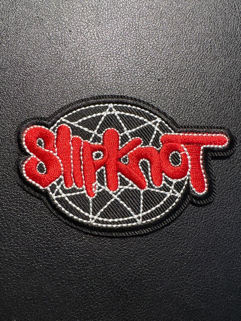 Może przedstawiać: Haftowana naszywka z napisem "Slipknot" w kolorze czerwonym, obramowanym na biało, na czarnym tle. Tekst jest w stylizowanej czcionce, z białym wzorem pajęczyny za tekstem. Naszywka ma czarną obw&oacute;dkę.